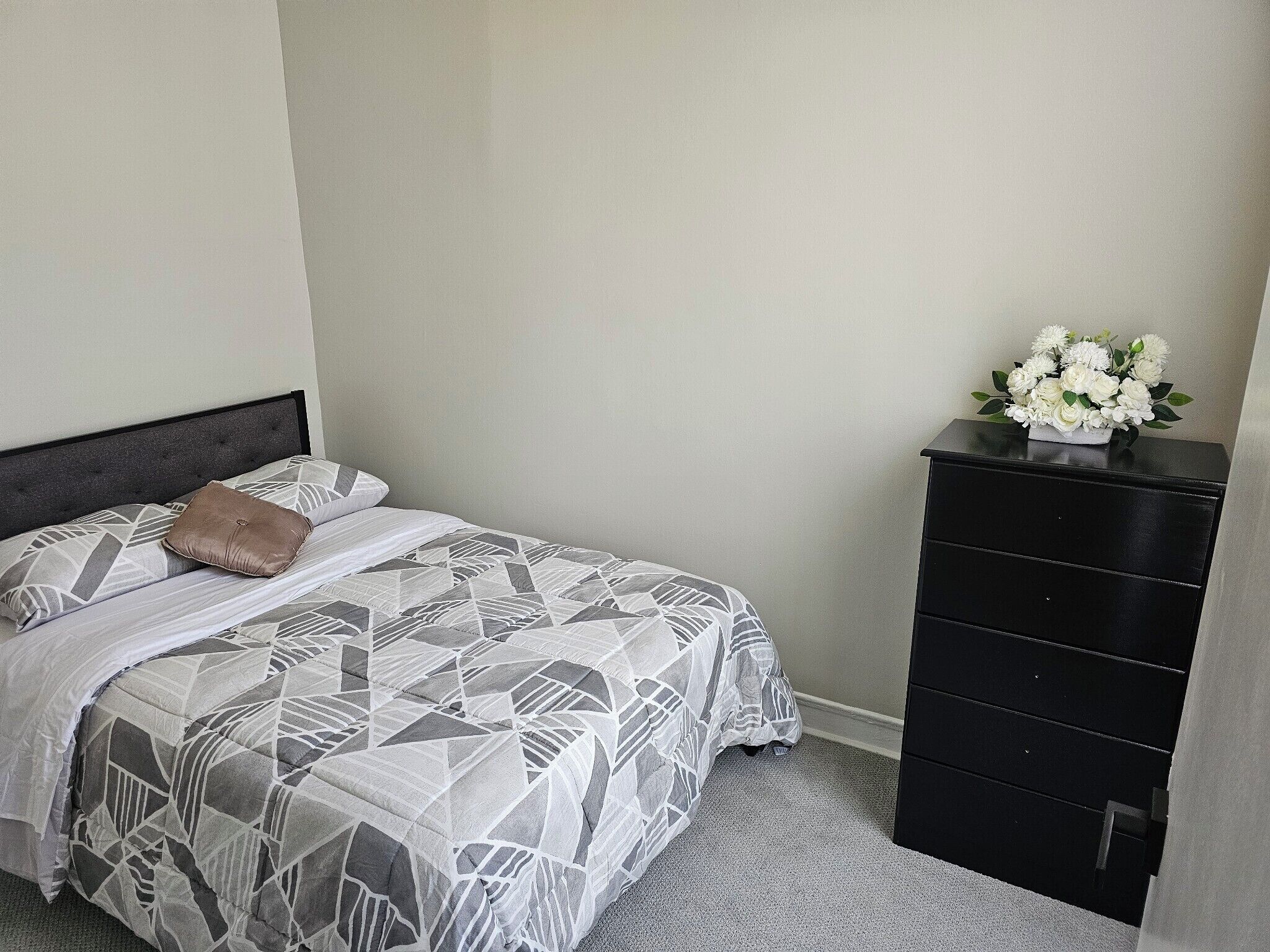 A neatly made bed with a geometric patterned bedspread and pillows, a black dresser with drawers, and a vase of white flowers on top.