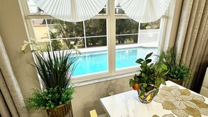 A room with a view of a swimming pool, a table with a decorative tablecloth, and potted plants. - Charming Lakeview Home, Heated Saltwater Pool, WiFi, All New Furniture (Venice)
