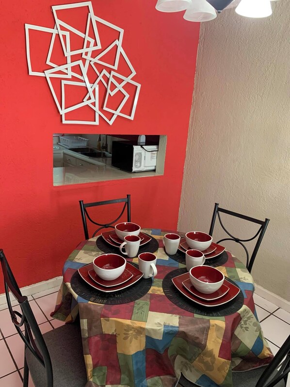 Un comedor con pared roja, un adorno mural geométrico, un espejo pequeño y una mesa dispuesta con cuencos y platos rojos y blancos.
