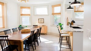 A modern kitchen with a large island, wooden dining table, and bar stools. There are large windows providing natural light and a view of the outdoors. - The Lounge- Heated Private Bar, Hot Tub, Fire Pit, Sleeps 14 (Cincinnati)