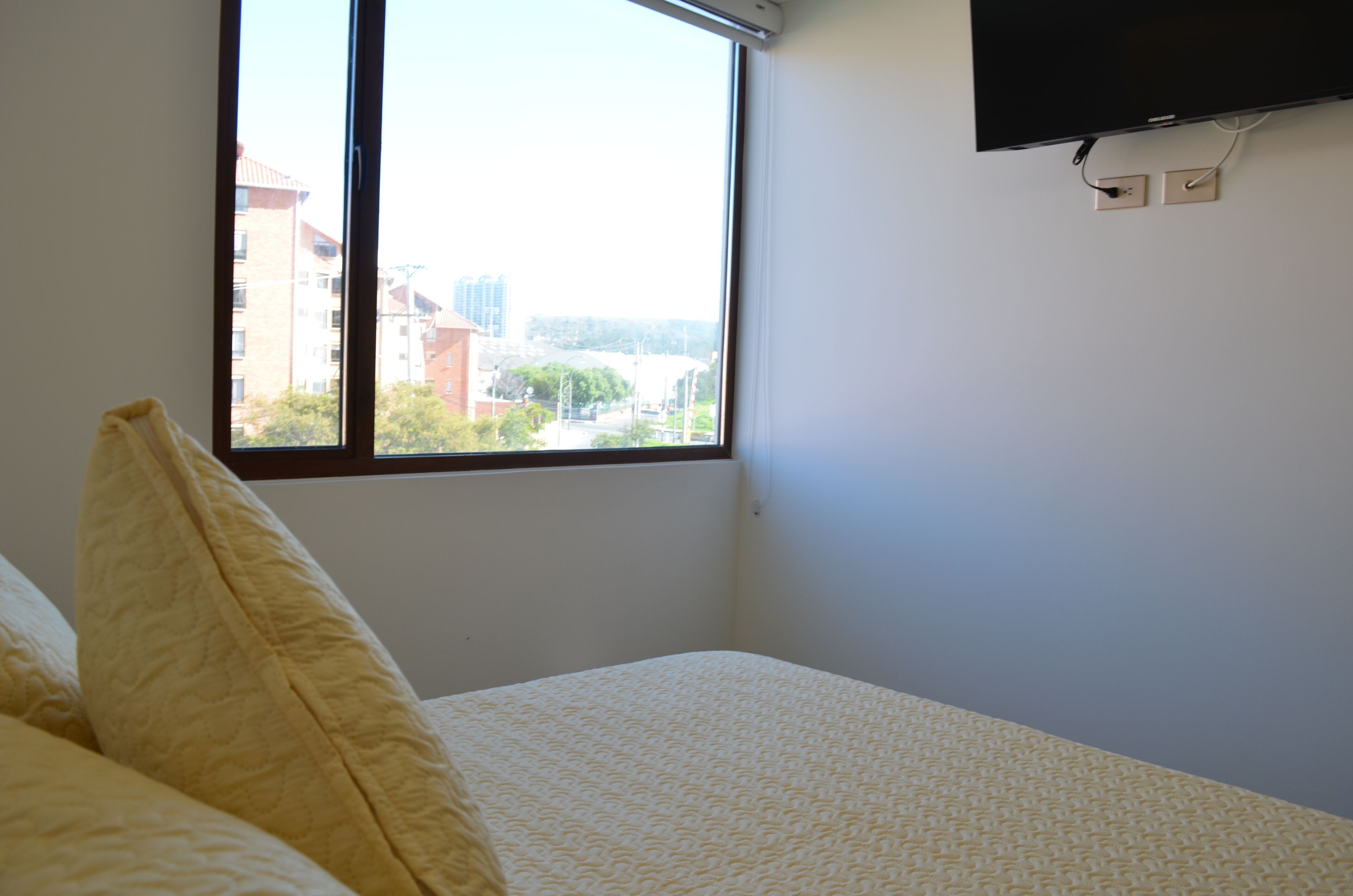 A hotel room with a bed, a window with a view of buildings, and a flat-screen TV mounted on the wall.