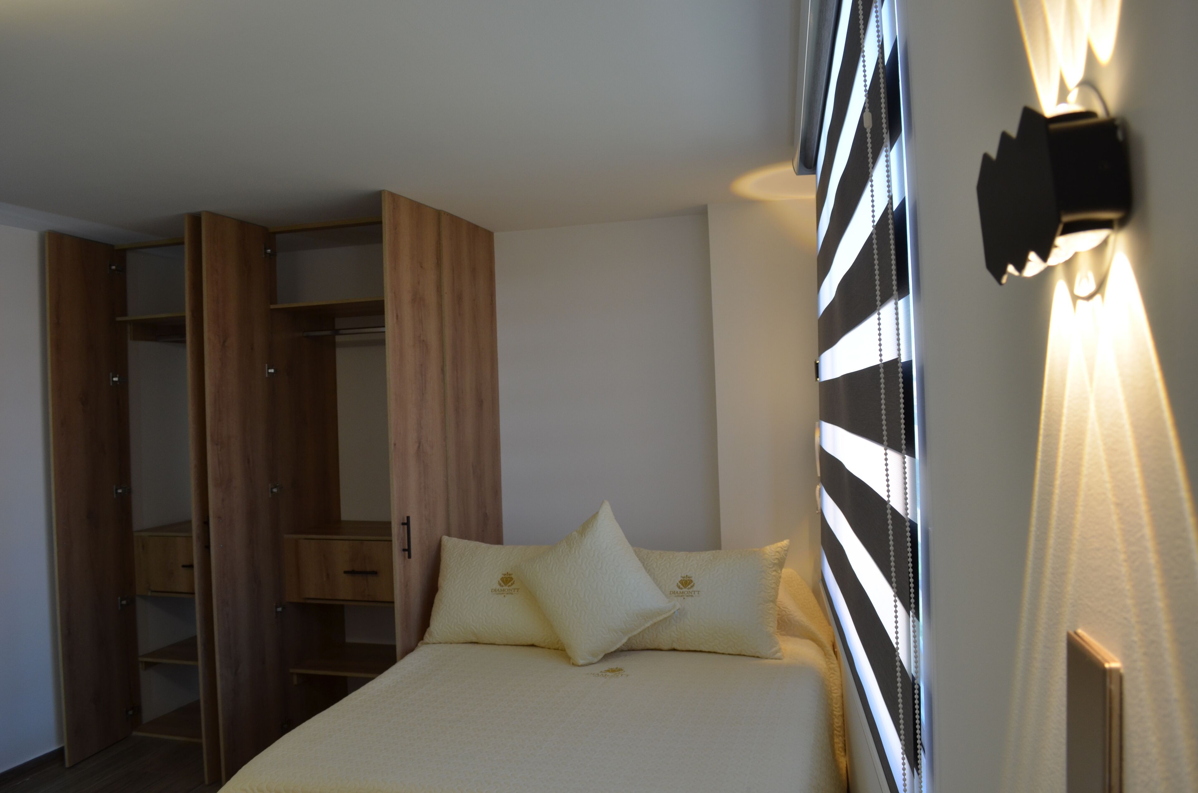 A bedroom with a bed, two pillows, a wooden wardrobe, and a wall-mounted light fixture.