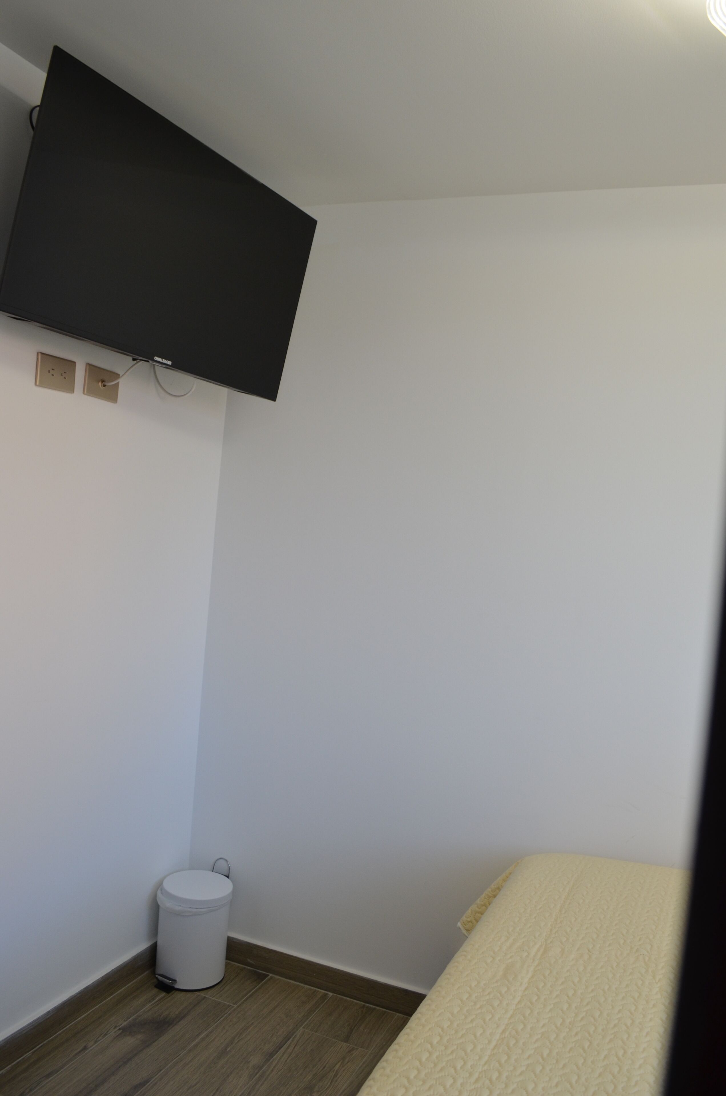 Una habitación pequeña con cama, televisor montado en la pared, bote de basura y toma de corriente.