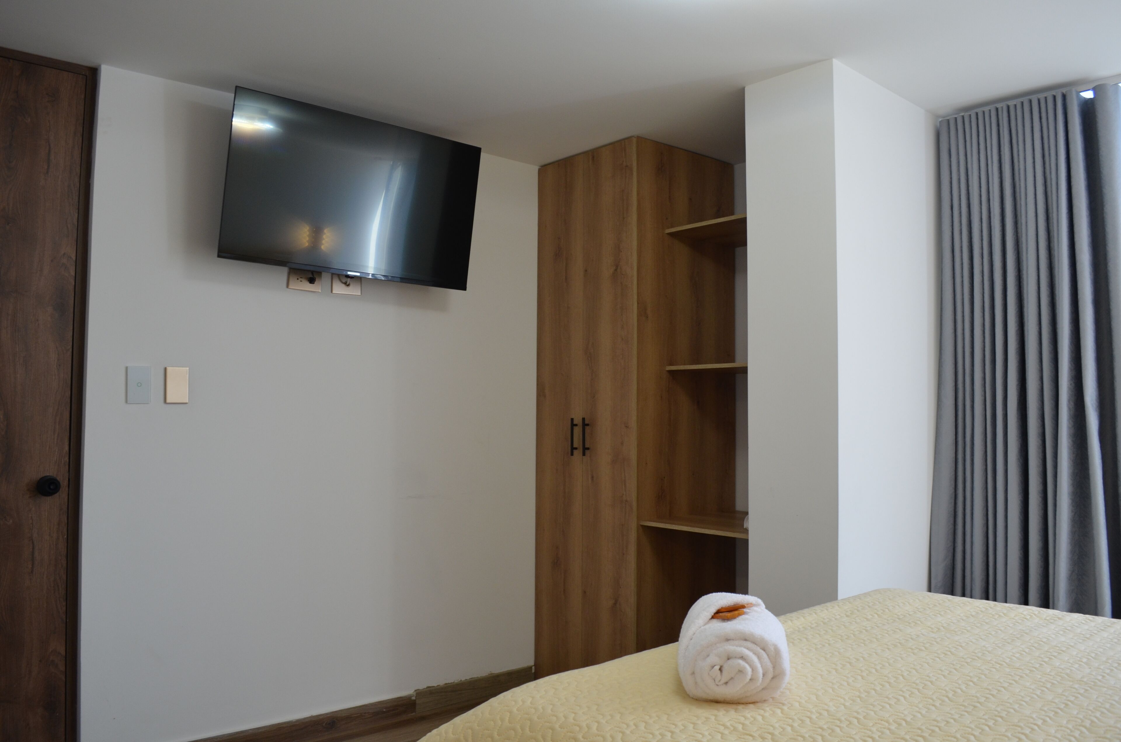 Una habitación de hotel con cama, televisión montada en la pared, armario de madera y cortina gris.