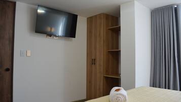 Una habitación de hotel con cama, televisión montada en la pared, armario de madera y cortina gris.