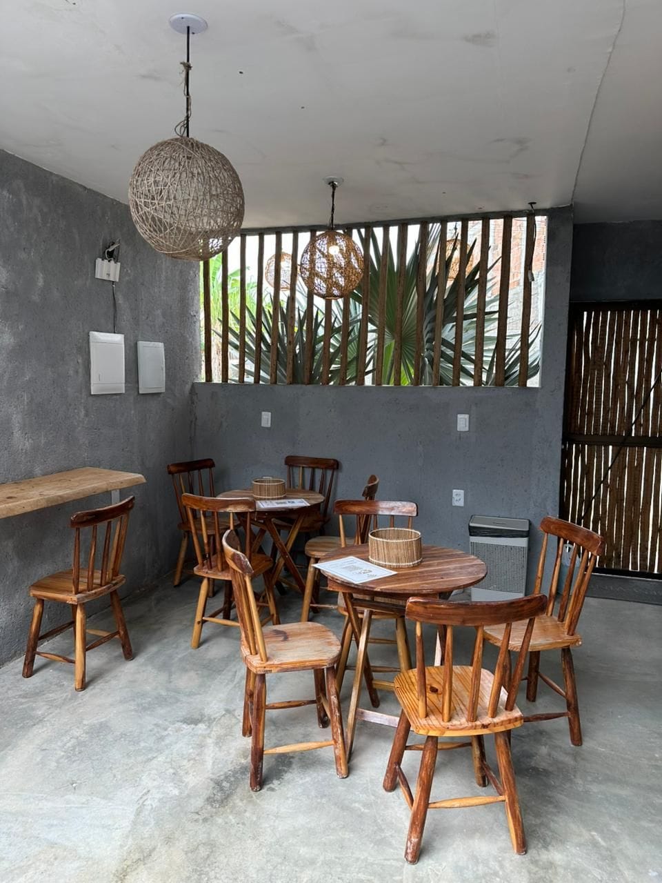 Un comedor con sillas y mesas de madera, una lámpara colgante y vistas a la vegetación a través de una ventana.