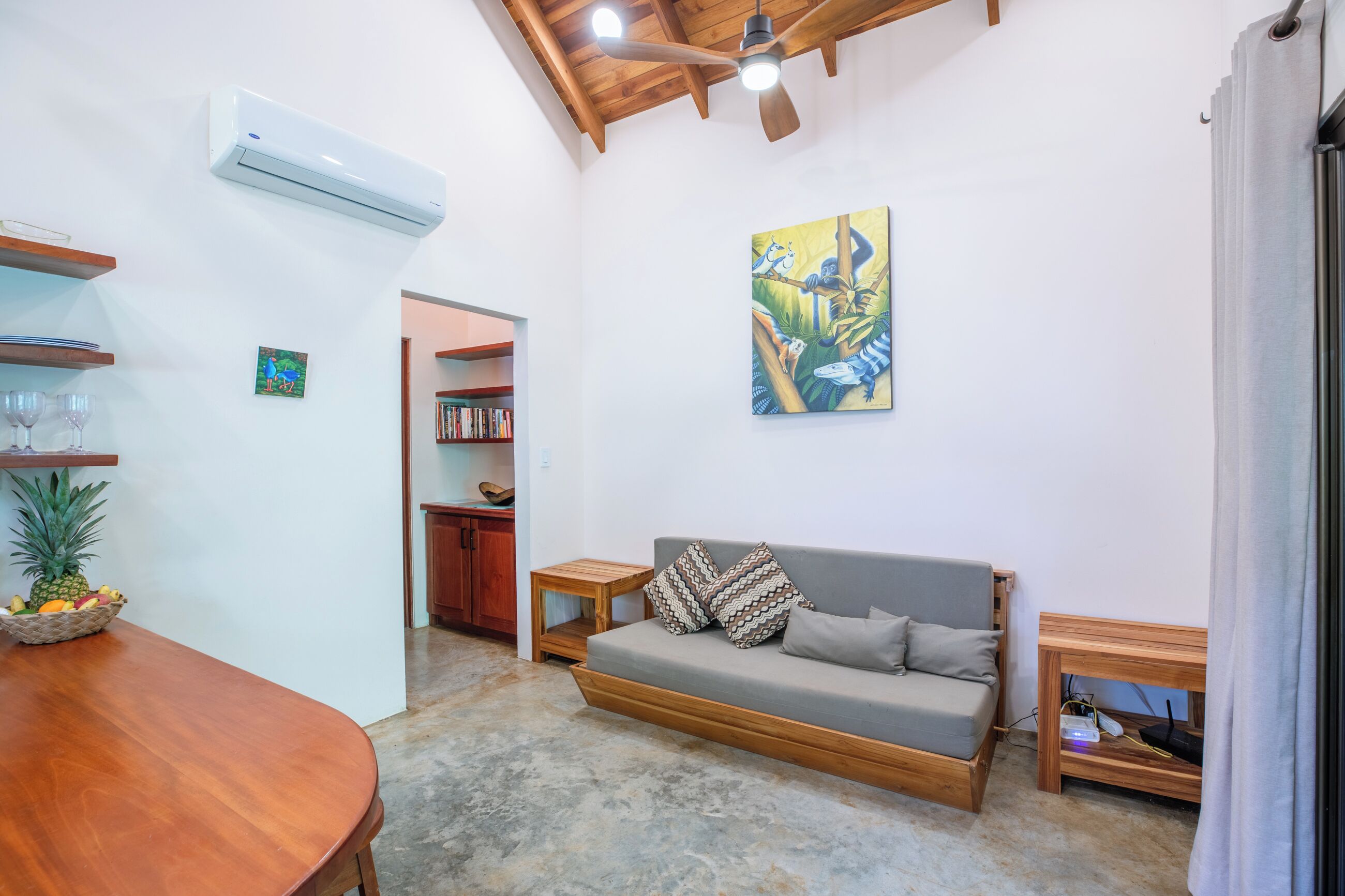 A living room with a sofa, a dining table with fruit, a ceiling fan, and a painting on the wall.
