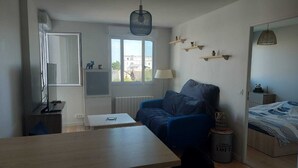 En kompakt opholdsstue med en sofa, et lille bord, en lampe, et fjernsyn og en seng med et mønstret dynebetræk.