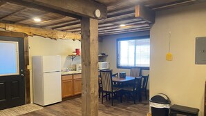 Cocina con refrigerador, microondas y gabinetes de madera. Hay una mesa de comedor con sillas y una ventana con persianas. También se ve un cubo de basura y un horno a leña.