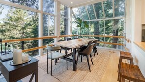 Un comedor con mesa y sillas de madera, ventanas amplias y vistas a árboles.