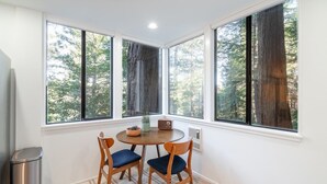 Habitación de esquina con una mesa redonda, dos sillas, un cesto de basura y ventanas grandes con vistas a árboles.