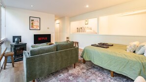 Una habitación acogedora con un sofá, una cama con colcha verde, una chimenea y un televisor de pared mostrando 'NETFLIX'.