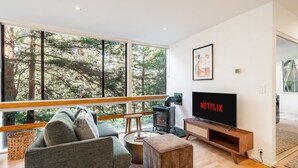 Una sala de estar moderna con un ventanal grande, chimenea, el logotipo de Netflix en la televisión y una zona de estar acogedora.
