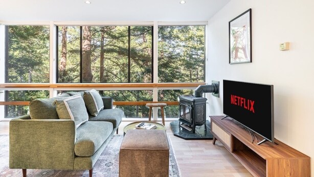 Una sala de estar moderna con un sofá, reposapiés y un televisor que muestra 'NETFLIX'. Se ve un estufa de leña y amplias ventanas ofrecen vistas a árboles.