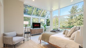 A modern bedroom with a large bed, a sofa, a TV on a wooden cabinet, and a view of trees through large windows.