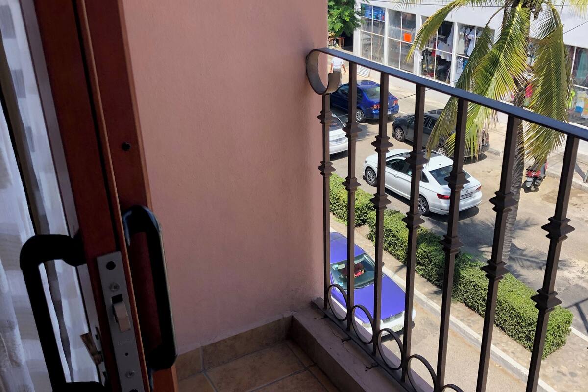 Een balkon met een metalen hekwerk, zicht op geparkeerde auto's en een gebouw met rode pilaren op de achtergrond.