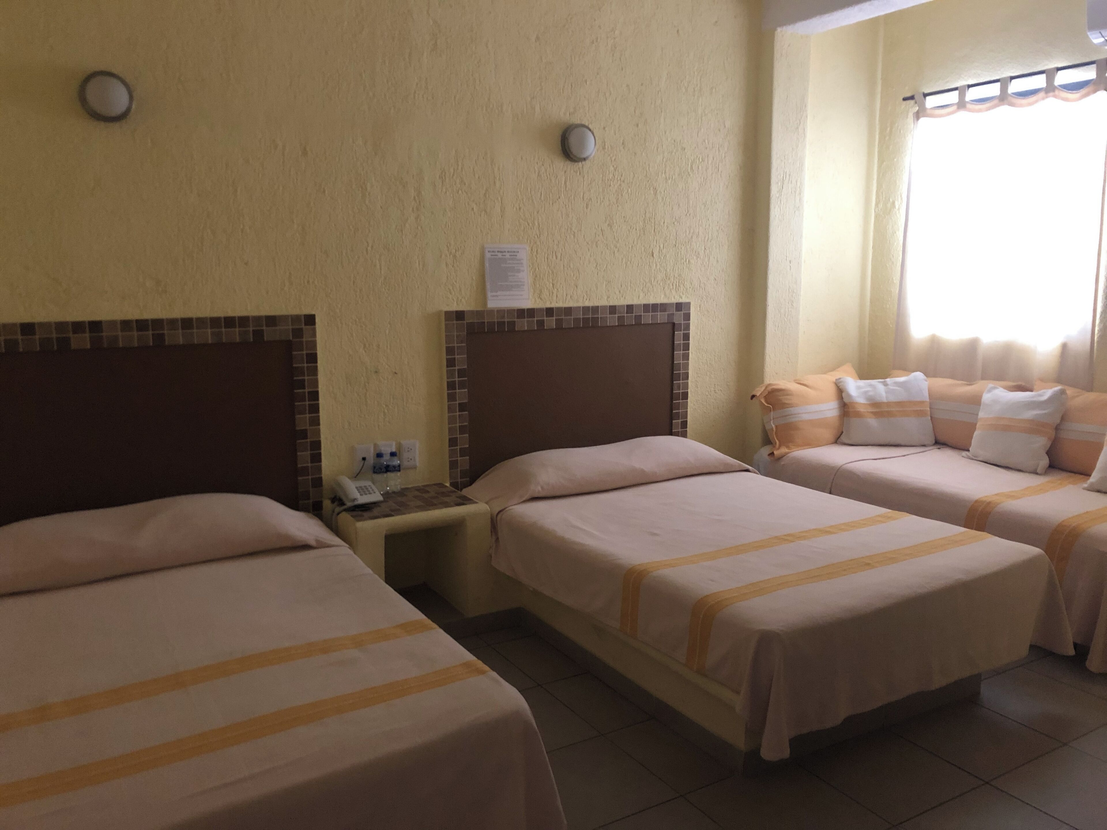 A hotel room with two single beds and a sofa bed, a small bedside table with a phone and water bottles, and a wall-mounted light fixture.