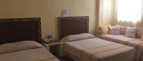 A hotel room with two single beds and a sofa bed, a small bedside table with a phone and water bottles, and a wall-mounted light fixture.