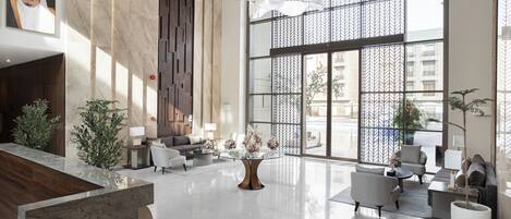 Un vestíbulo amplio con un mostrador de recepción de mármol, asientos modernos, ventanas grandes y una pared decorativa.