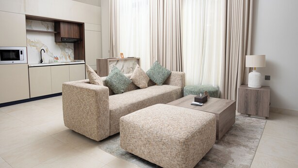 A modern living room with a sofa, ottoman, and a coffee table. There is a kitchen area with a microwave and cabinets. - Liora Al Yasmin District  (Riyadh)