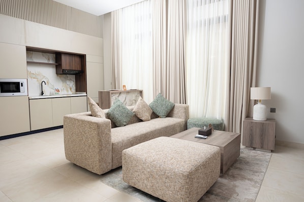 Una sala de estar moderna con un sofá, un reposapiés y una mesa de centro. Hay una zona de cocina con microondas y armarios.