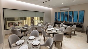 A modern dining area with multiple tables and chairs, a large window with artwork, and an air conditioning unit on the wall. - Liora Hotel (Riyadh)