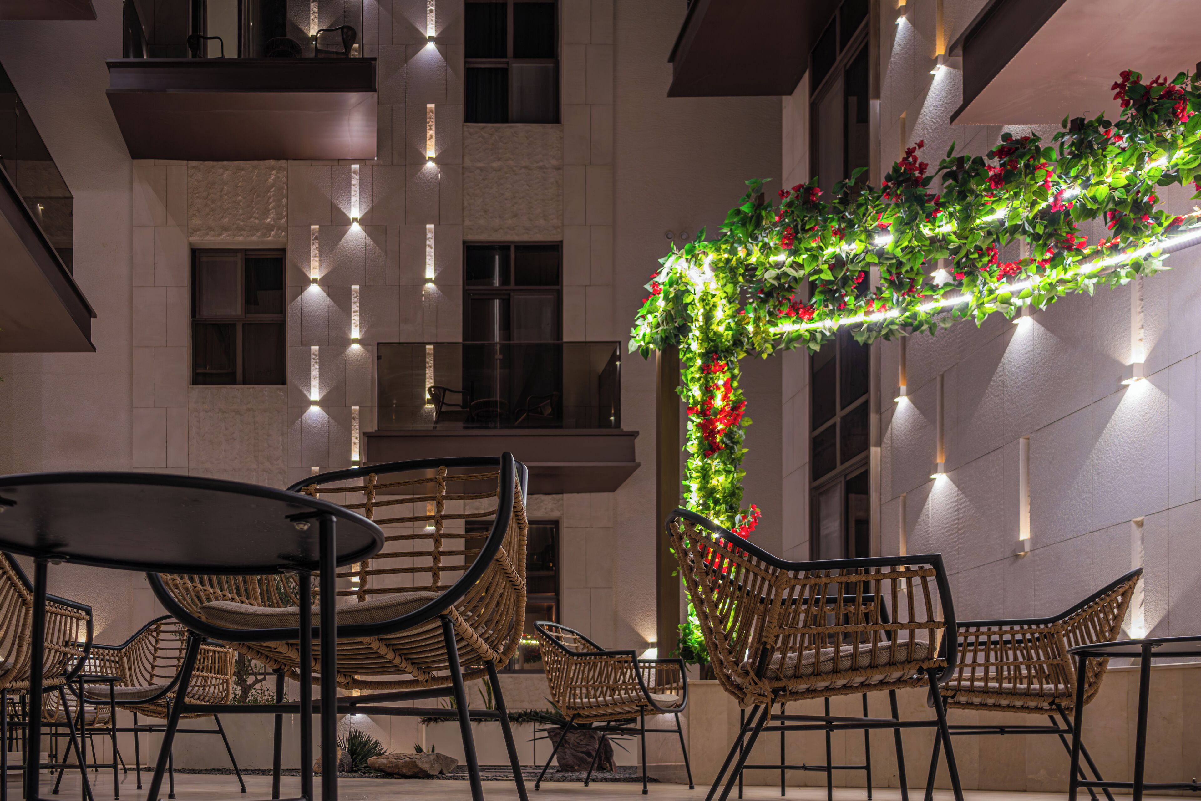An outdoor seating area with wicker chairs and tables, a decorative arch with greenery and lights, and a building facade with windows and light fixtures.