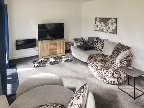 A living room with a sofa, a coffee table, a flat-screen TV on a wooden cabinet, and a floral painting on the wall. - Classroom Two - UK48651 (Lifton)