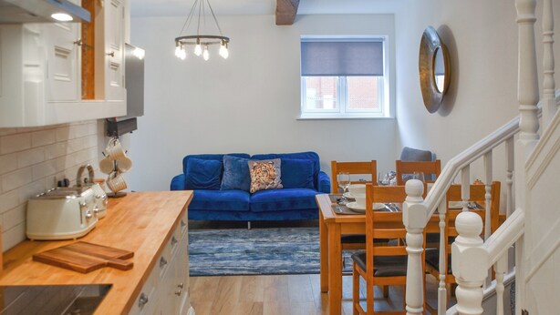 A compact kitchen with modern appliances, a dining area with wooden furniture, a blue sofa, and a staircase with white balusters. - Norfolk Headquarters (Fakenham)