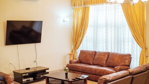 A living room with a brown leather sofa, a coffee table, and a flat-screen TV mounted on the wall. - Eey Kay Booking Ghana (Accra)