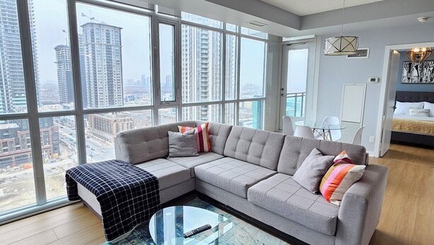 A modern living room with a sectional sofa, a glass coffee table, and a large window offering a city view. - Beautiful Condo with spectacular views + 1 Parking in Mississauga Downtown (Mississauga)
