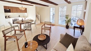 Ein geräumiges Wohnzimmer mit einer Holzbank, einem runden Tisch mit einer Flasche und Gläsern sowie einem Sofa mit Kissen. Eine Treppe führt zu einer Küche mit Holzausstattung und einem Essbereich.