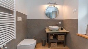 A modern bathroom with a white toilet, a round mirror with a light above it, a white sink with a soap dispenser, and a wooden shelf with baskets. - Ciesa dela Maestra 2ªB (Rocca Pietore)