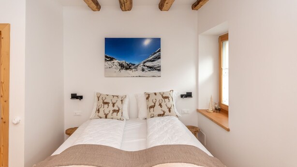A bedroom with a bed, wooden beams, a mountain landscape painting, and a small shelf with a model tree. - Ciesa dela Maestra 2ªB (Rocca Pietore)