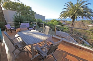 An outdoor patio with a wooden table and chairs, surrounded by a railing, overlooking a scenic landscape with trees and a view of the sea.
