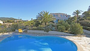 A swimming pool with a stone deck, surrounded by lush greenery and a hillside in the background.