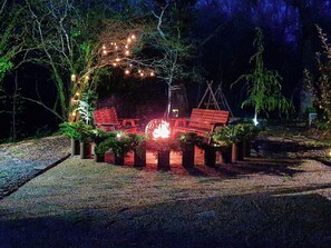 Un espacio al aire libre con mesas y sillas de madera, bancos y luces cálidas colgantes.