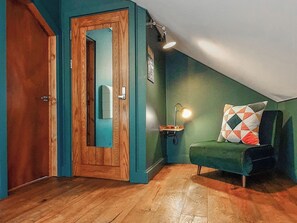 Una habitación con puerta de madera, un espejo, pared verde, una mesa pequeña con lámpara y una silla verde con cojín estampado.