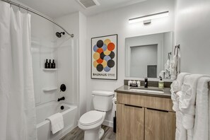 Un baño moderno con bañera, inodoro y lavabo. Tiene una cortina de ducha, una obra de arte enmarcada en la pared y un perchero con una toalla.
