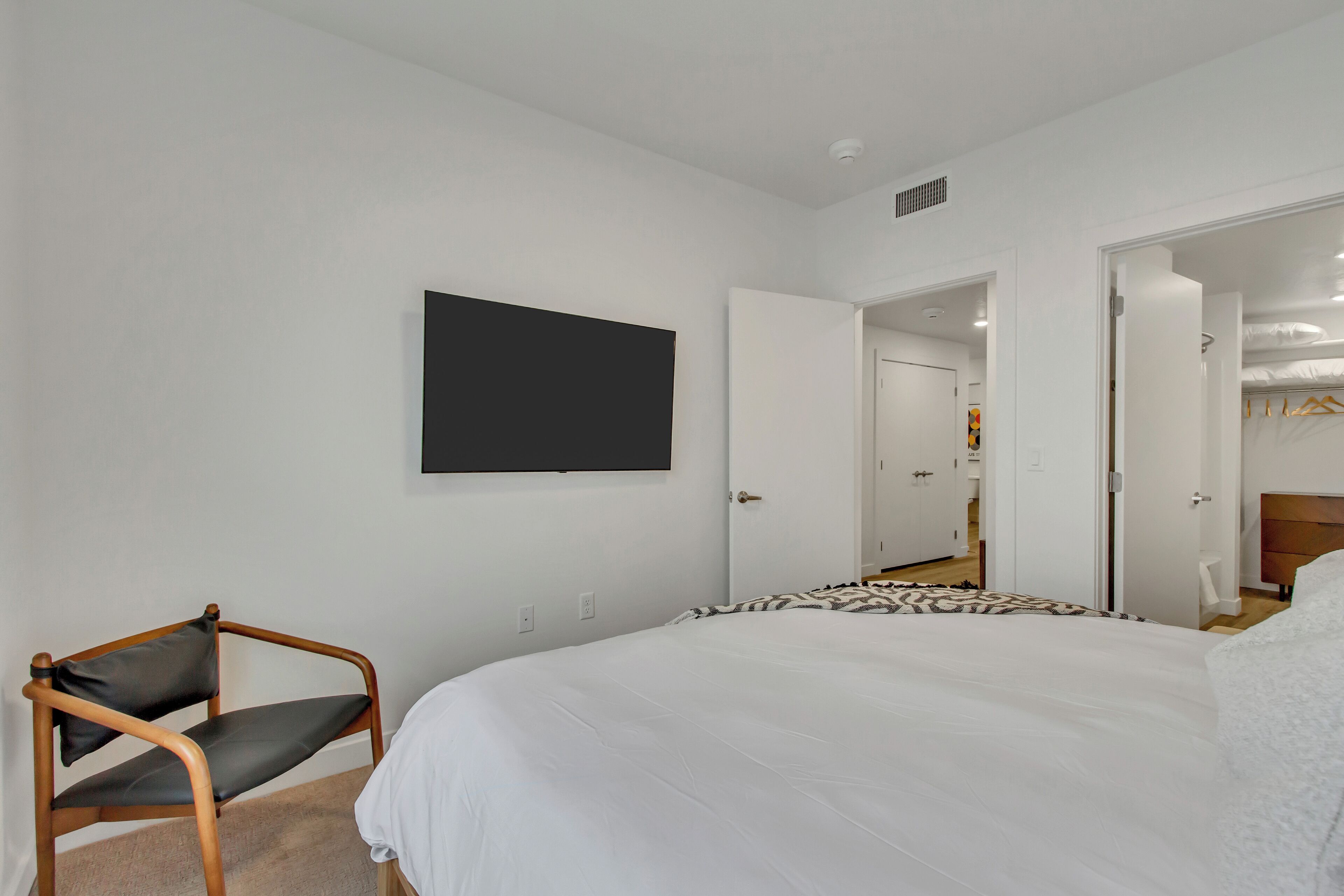 Un dormitorio con una cama, una silla, un televisor montado en la pared y una puerta abierta que da a otra habitación.