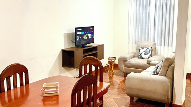 Een woonkamer met een houten eettafel, stoelen, een bank, een televisie op een meubelstuk en een klok aan de muur.