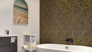 A bathroom with a white bathtub, a toilet, and a vanity with a mirror. There is a painting of a beach scene on the wall, and a patterned wallpaper. - Classroom One - UK48650 (Lifton)