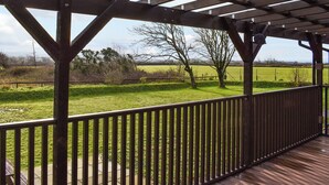 A covered outdoor area with a wooden deck and metal railing, overlooking a grassy field and trees. - Classroom One - UK48650 (Lifton)