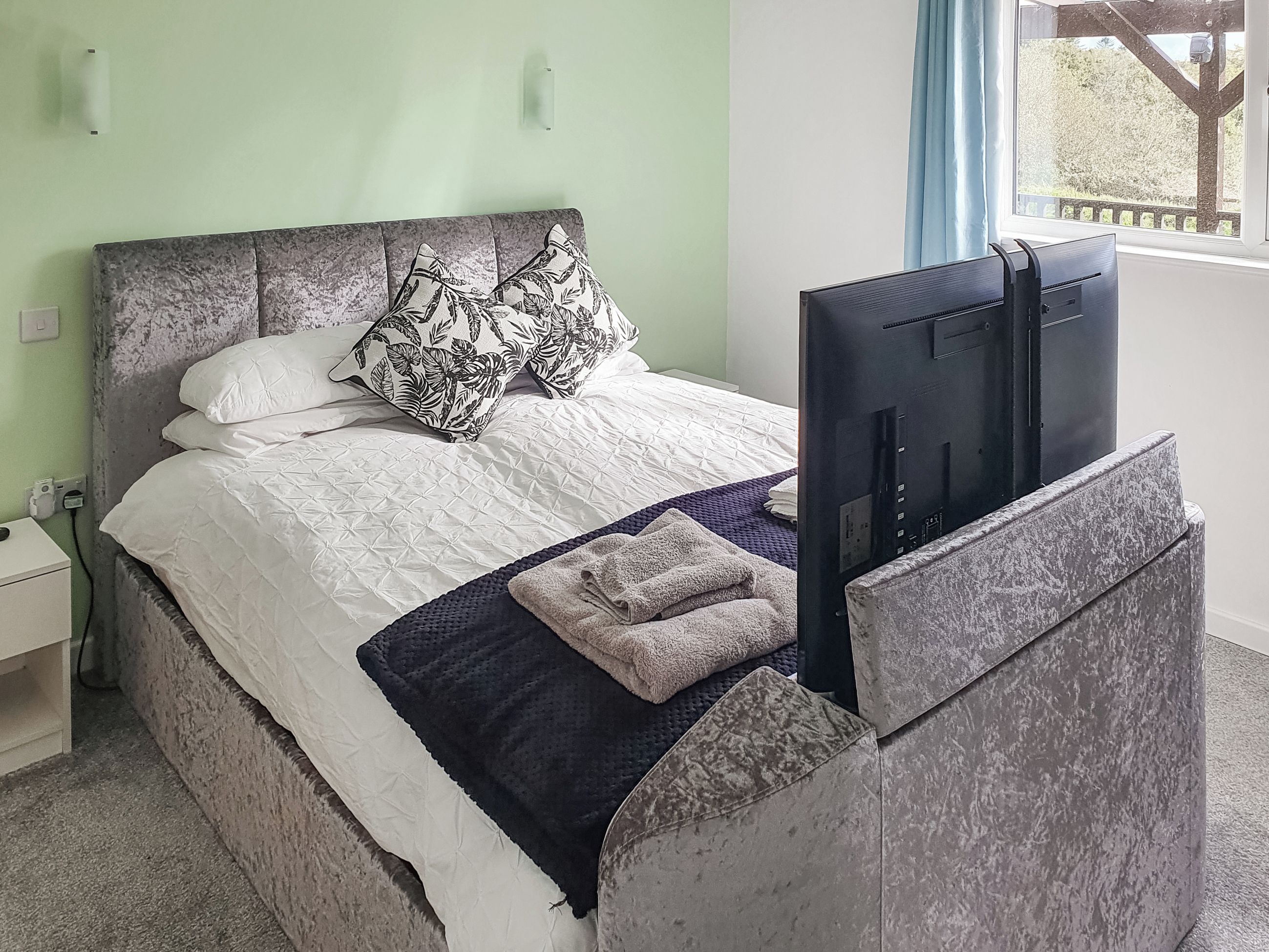 Een bed met een grijs hoofdeinde, witte beddengoed en een zwart-wit gestreept kussen. Een flatscreen televisie is aan het hoofdeinde bevestigd.