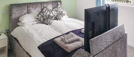 A bed with a grey headboard, white bedding, and a black and white patterned pillow. A flat-screen TV is mounted on the headboard.