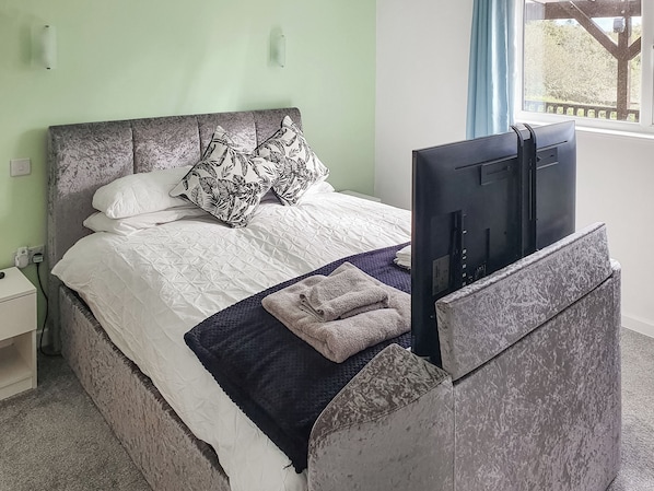 A bed with a grey headboard, white bedding, and a black and white patterned pillow. A flat-screen TV is mounted on the headboard.
