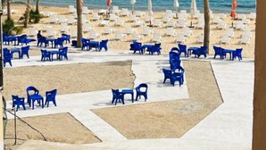 Una zona frente a la playa con hileras de sillas y mesas de plástico azul, sombrillas blancas y palmeras.