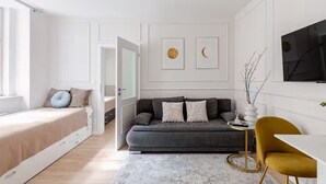 A modern living room with a grey sofa, a white coffee table, and a yellow armchair. There is a bed with a beige headboard and a window with white curtains.