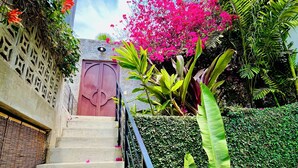 Un escalier menant à un bâtiment doté d’une porte rose, entouré d’une végétation luxuriante et de fleurs roses.