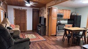 Un salon chaleureux avec un canapé, une table à manger avec des chaises, un coin cuisine doté de meubles en bois et d’appareils électroménagers, et un tapis à motifs sur le sol.